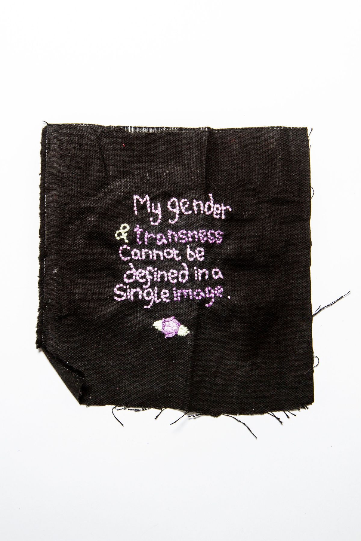 Piece of black cotton fabric with embroidered text in centre, square, frayed on all edges & unhemmed. Embroidered with mauve, purple & light green thread, reads; 'My gender & transness cannot be defined in a single image' with flower & leaves motif beneath