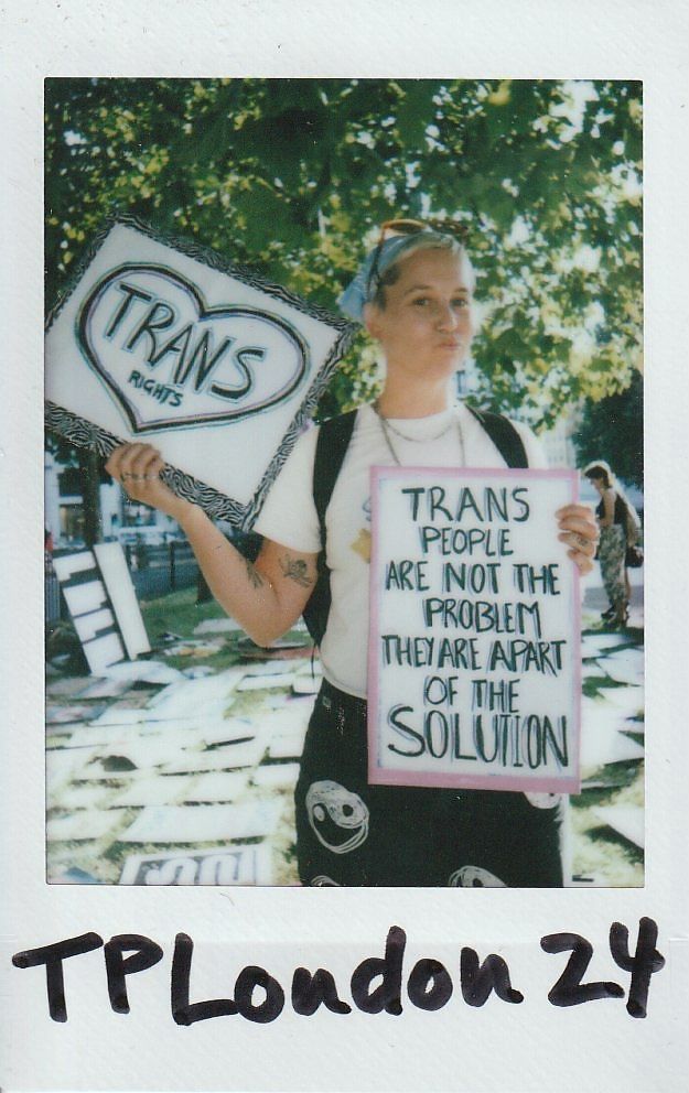 A person holds two signs advocating trans rights, standing outside under trees with sunlight filtering through leaves.