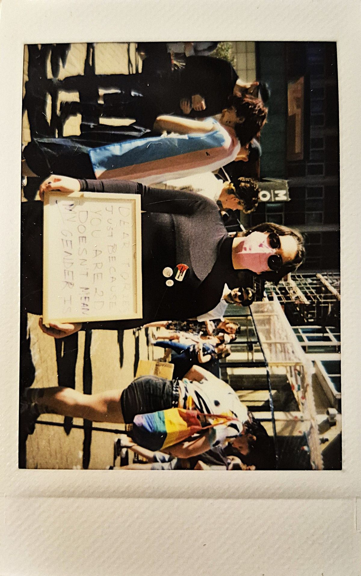 A person wearing a mask holds a sign at a busy outdoor protest or gathering event.