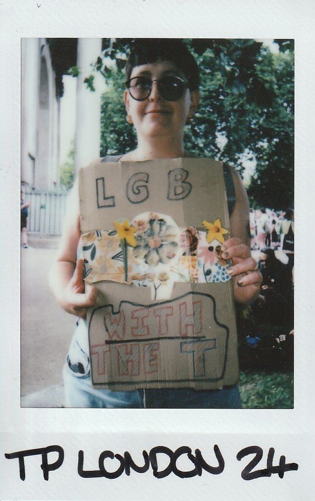 A person holds a cardboard sign reading "LGB with the T," decorated with flowers and stars.