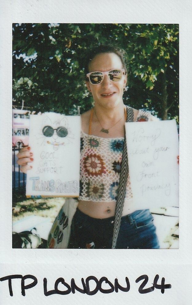 A person wearing sunglasses holds two signs advocating for trans rights while standing outdoors by trees.