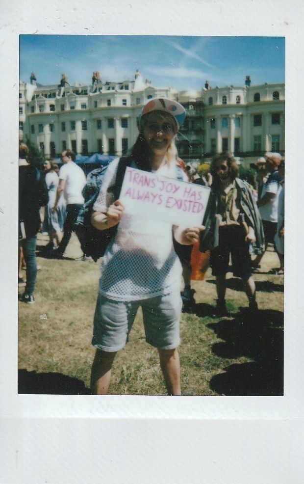 A person stands outside holding a sign that says "Trans Joy Has Always Existed" in bright sunlight.