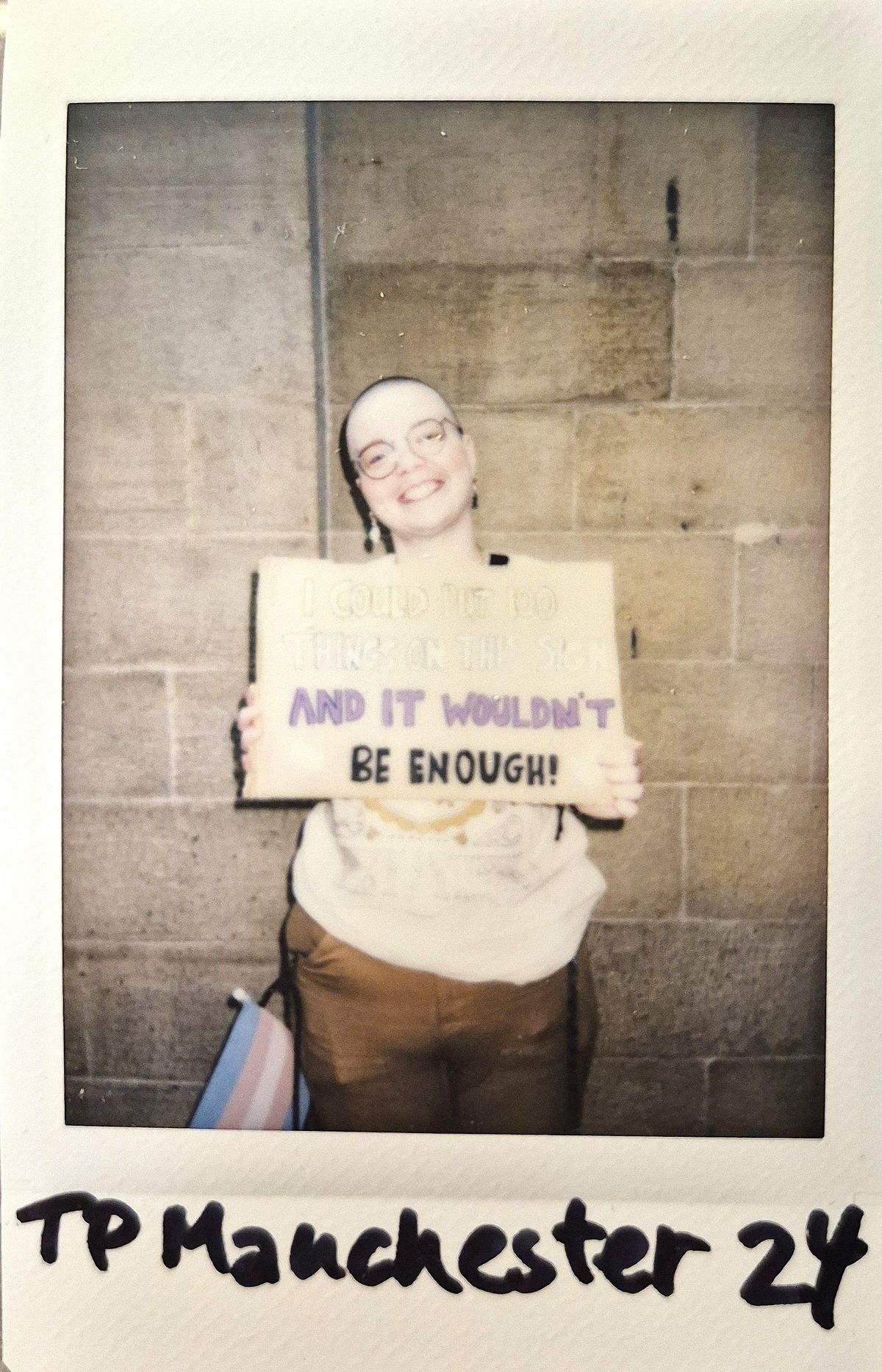 A person stands smiling against a stone wall, holding a placard.