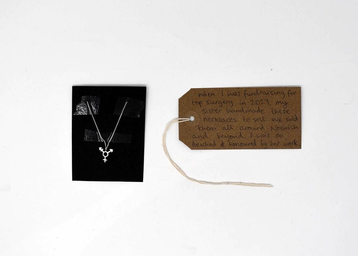 A necklace with the trans symbol displayed in a box with a brown luggage tag next to it.