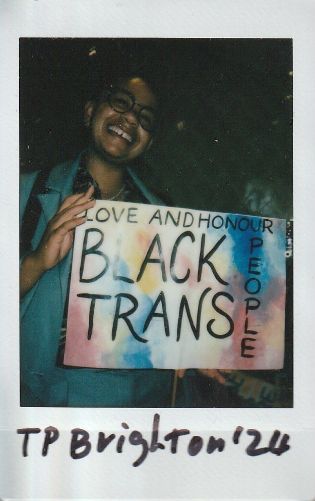 A person smiles while holding a colorful sign that says, "Love and Honour Black Trans People."