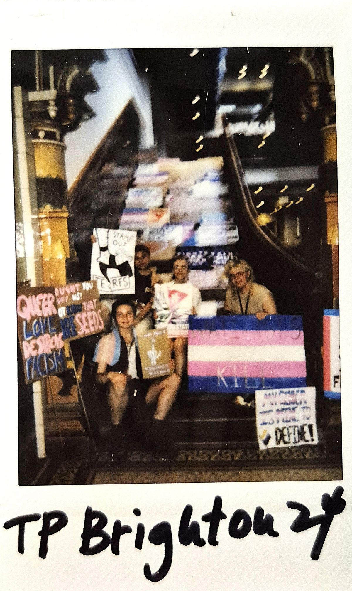 A group of people sits on stairs holding signs with messages supporting LGBTQ+ and social causes.