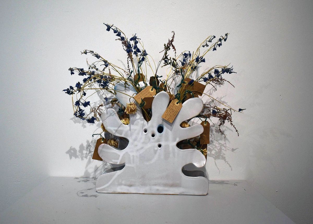 A splatter or blob shaped white ceramic vase holding dried flowers with brown, handwritten tags.