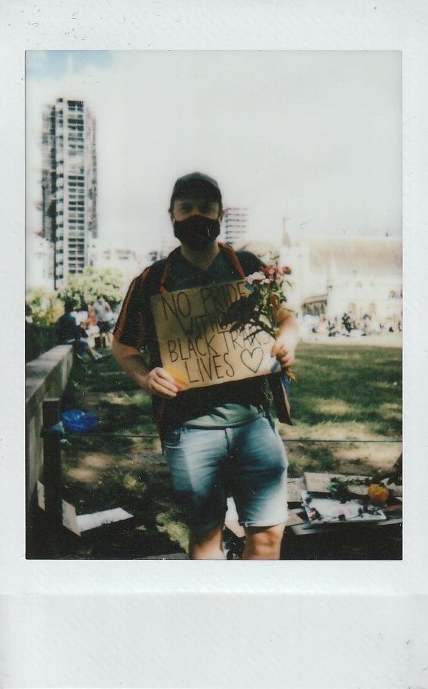 A person, wearing a mask, holds a sign which says "No Pride Without Black Trans Lives" standing in a park.