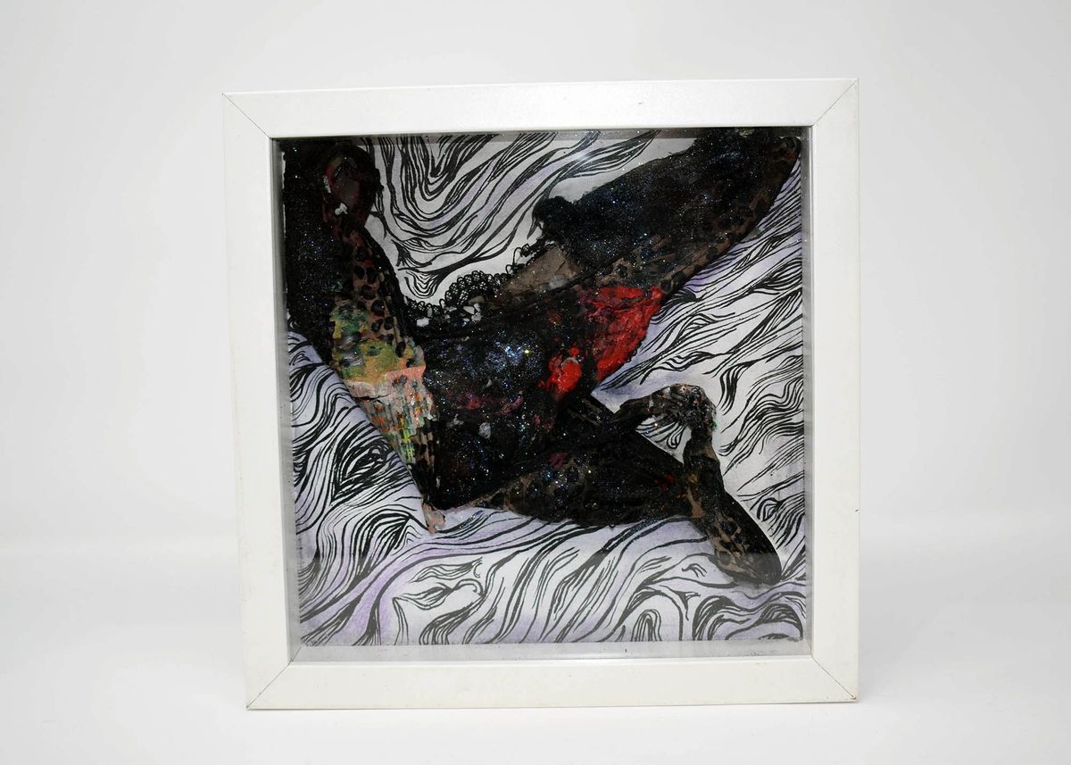 Black underwear covered in various paints and inks mounted on a white background in a white frame. The background has black ink lines drawn horizontally across the top, and swooping round lines moving in loops from the bottom edges, to the center, and back to the edges again.