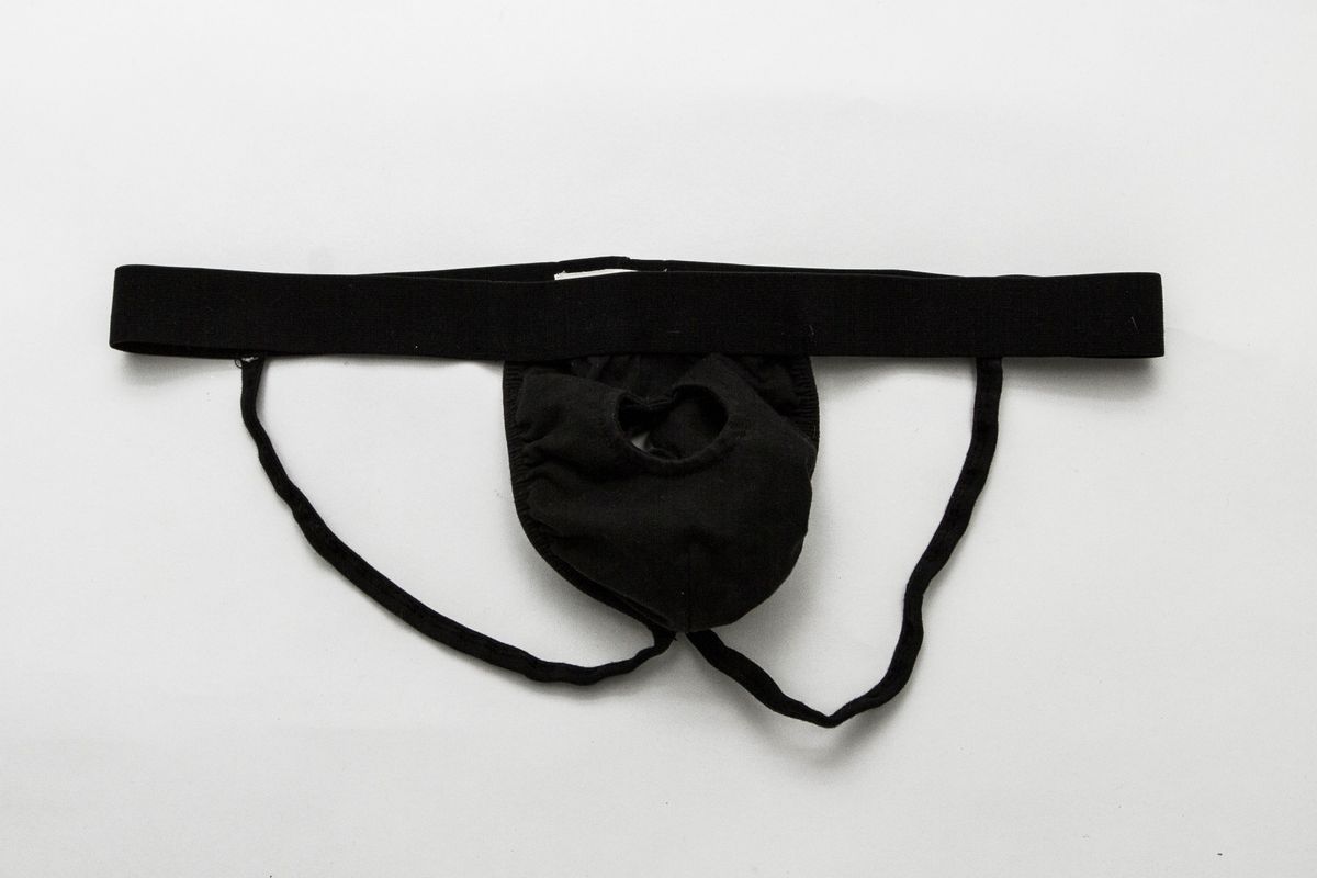 A plain black jockstrap.The elasticated waistband contains a white brand label. The front sack of the jockstrap has two layers, with the outer layer showing a round hole with a stitched edge.