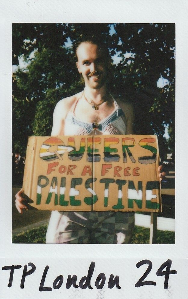 A person stands outdoors holding a sign reading "Queers for a Free Palestine" in colorful letters.