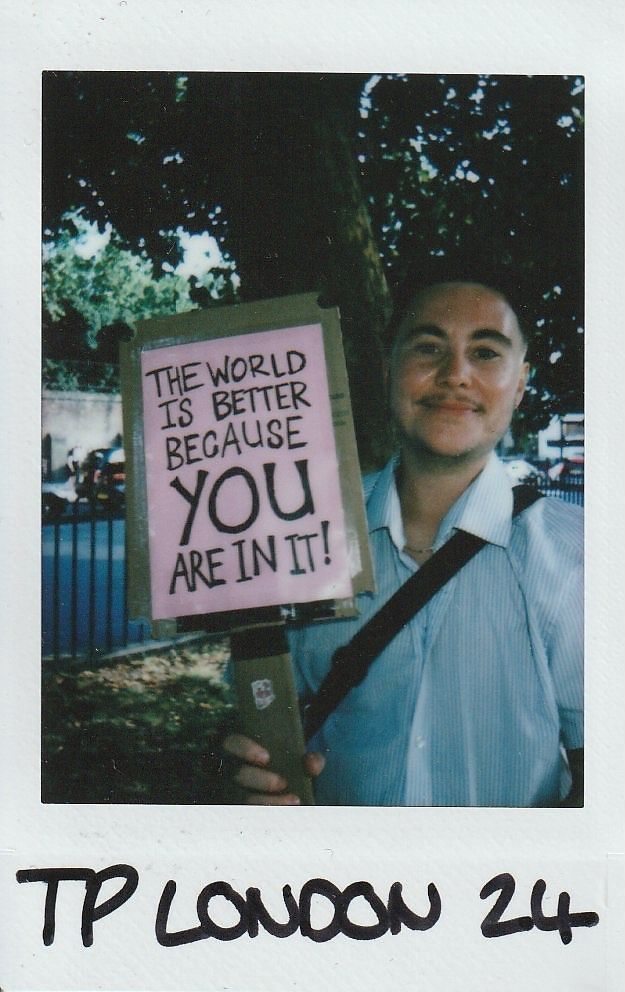 A person stands outside, smiling and holding a sign that reads, "The world is better because you are in it!"