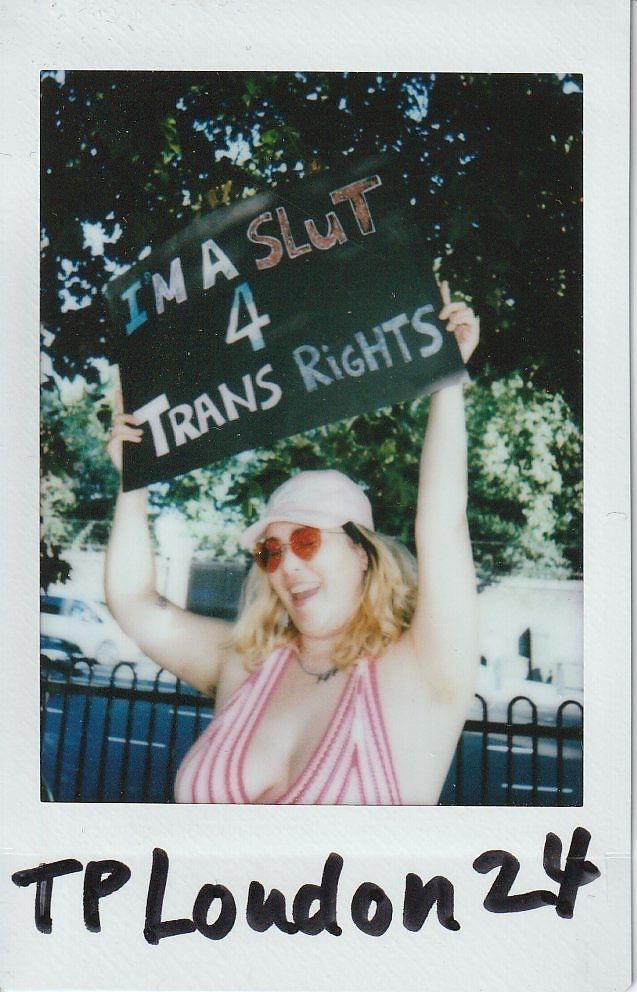 A person holds a sign supporting trans rights while smiling outdoors, wearing sunglasses and a striped top.