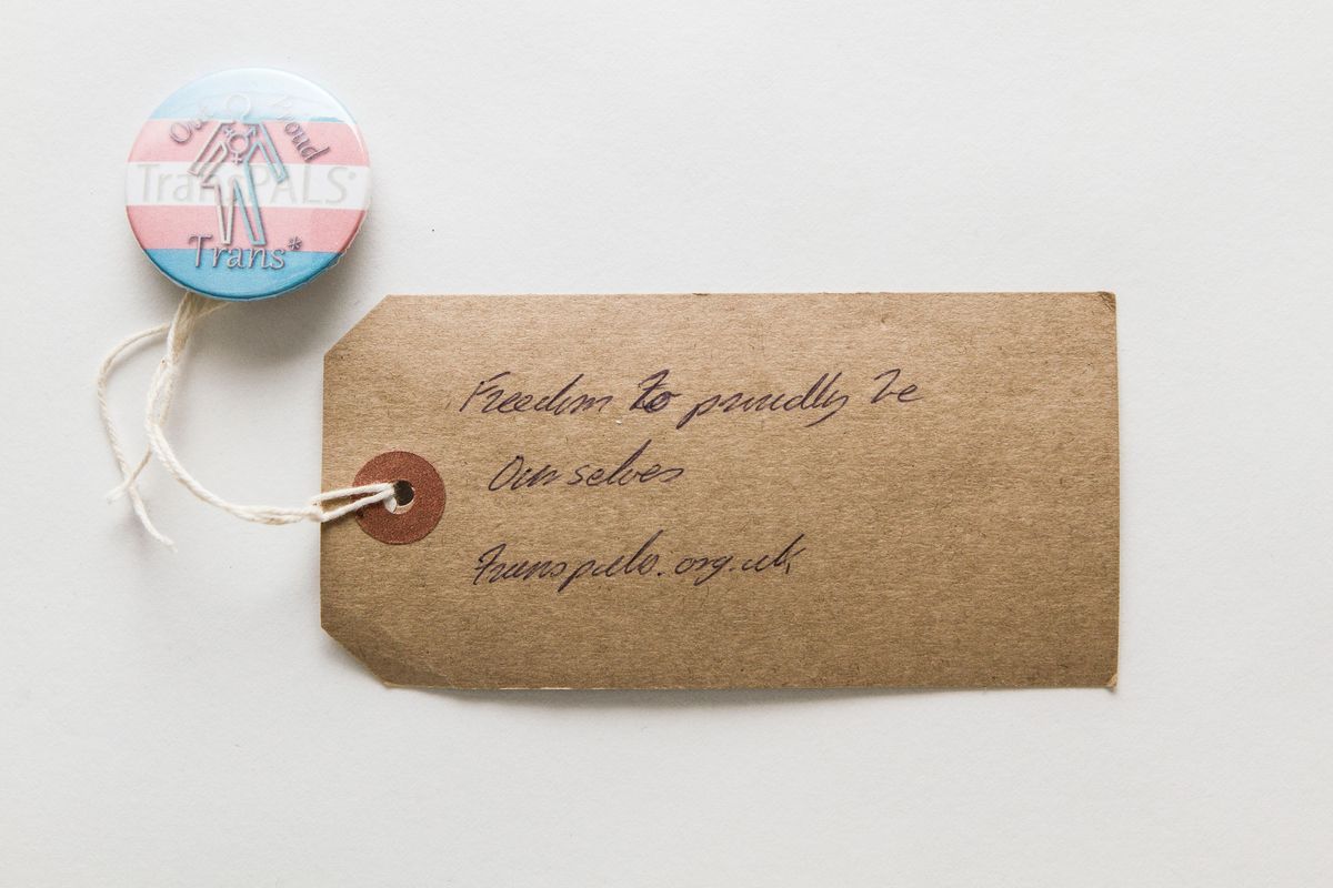 A badge which says 'Out And Proud' with a trans flag as the background and a figure.