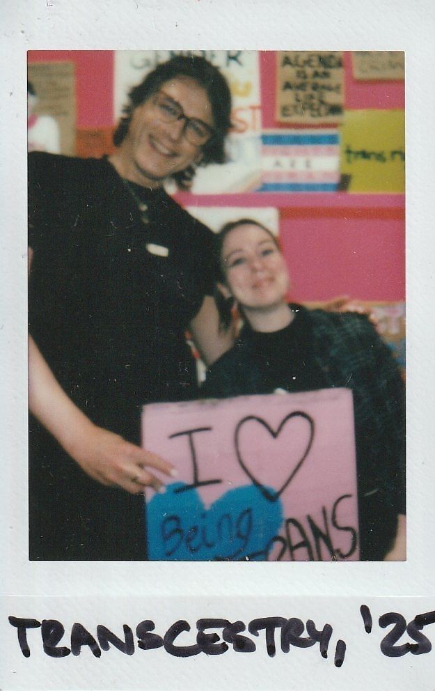 Two people smiling happily hold a pink sign that reads, "I love being trans," against a colorful background.