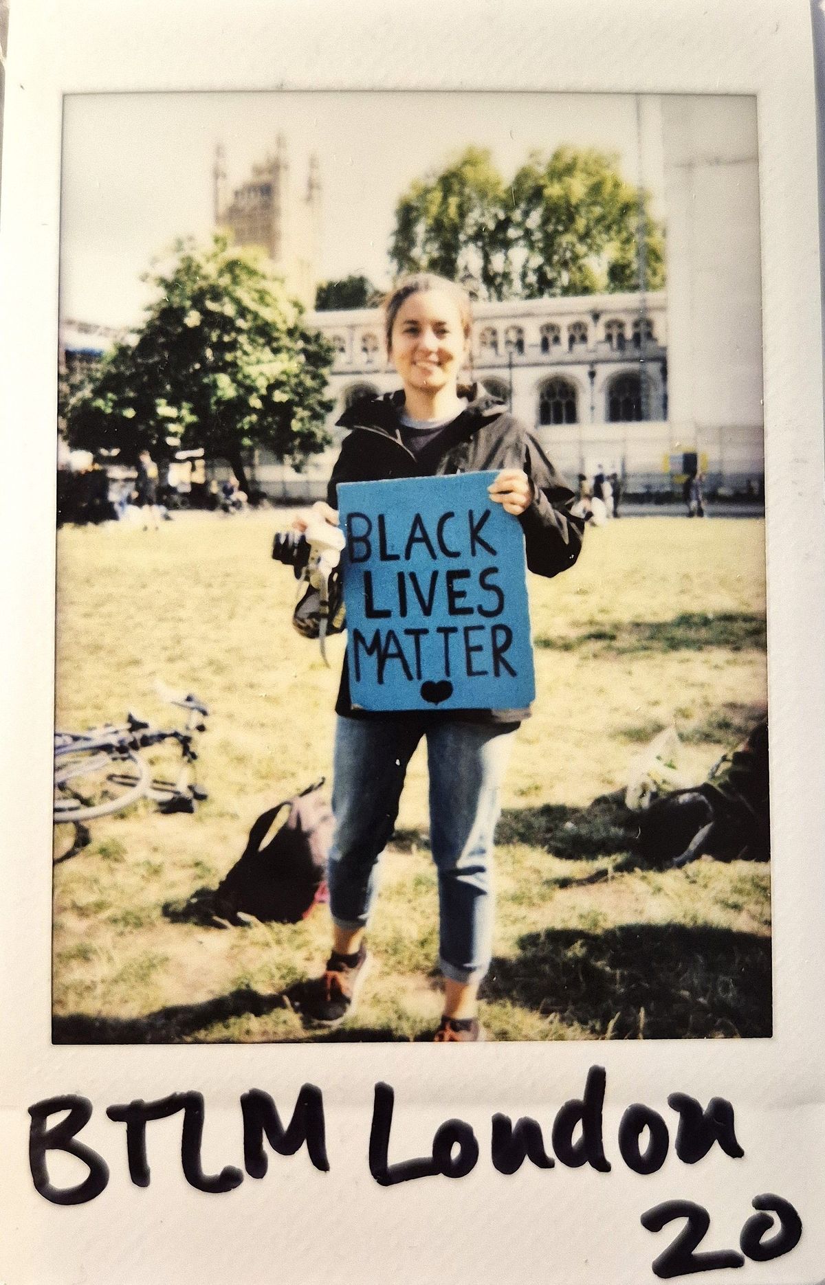 A person is holding a "Black Lives Matter" sign outdoors near a historic building and trees.