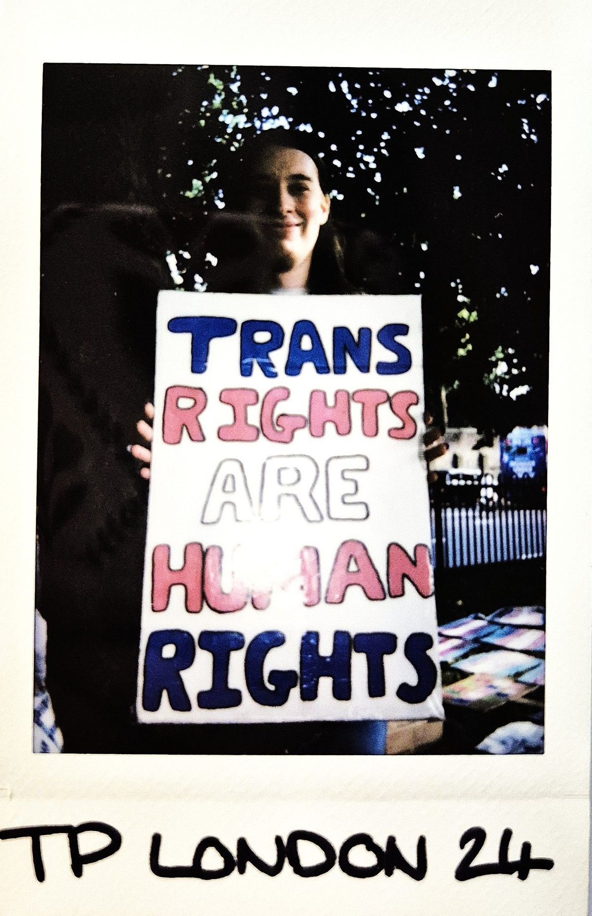 A person holds a sign reading "TRANS RIGHTS ARE HUMAN RIGHTS," with trees in the background.