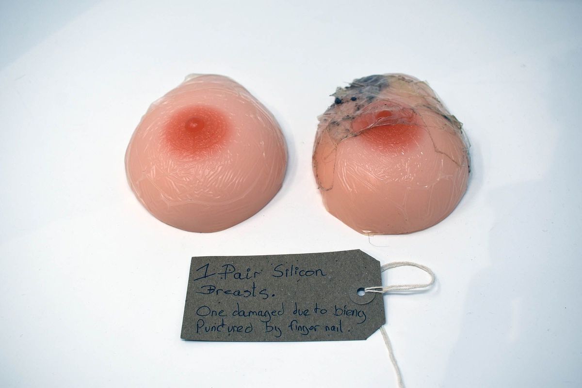 The image shows a pair of silicone breasts, one damaged, with a handwritten tag describing them.