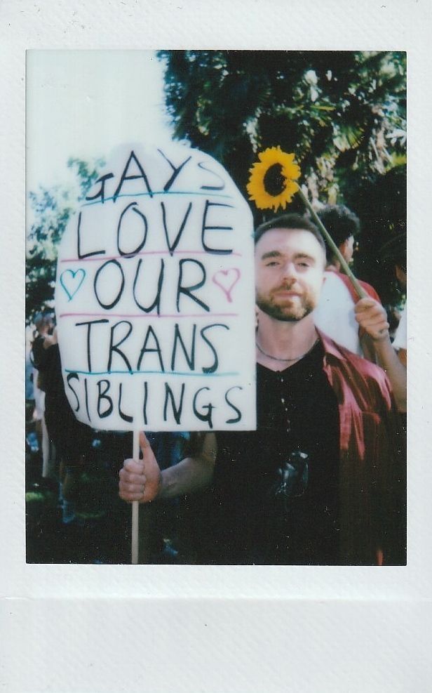 A person at a rally holds a sign saying "GAYS LOVE OUR TRANS SIBLINGS".