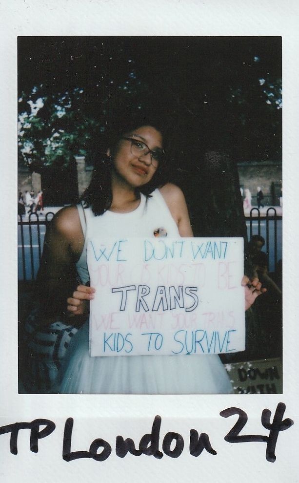 A person holds a sign which says "We Don't Want Your Cis Kids To Be Trans We Want Your Trans Kids To Survive" standing outdoors, near trees, wearing glasses and a white dress.
