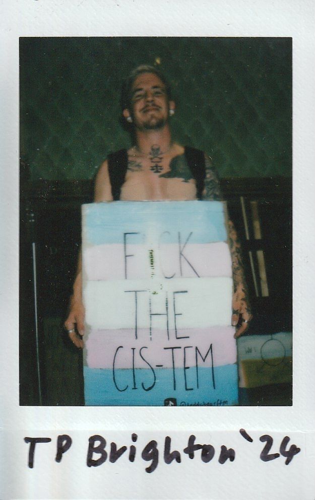A person with tattoos is holding a sign with text in a dimly lit room.
