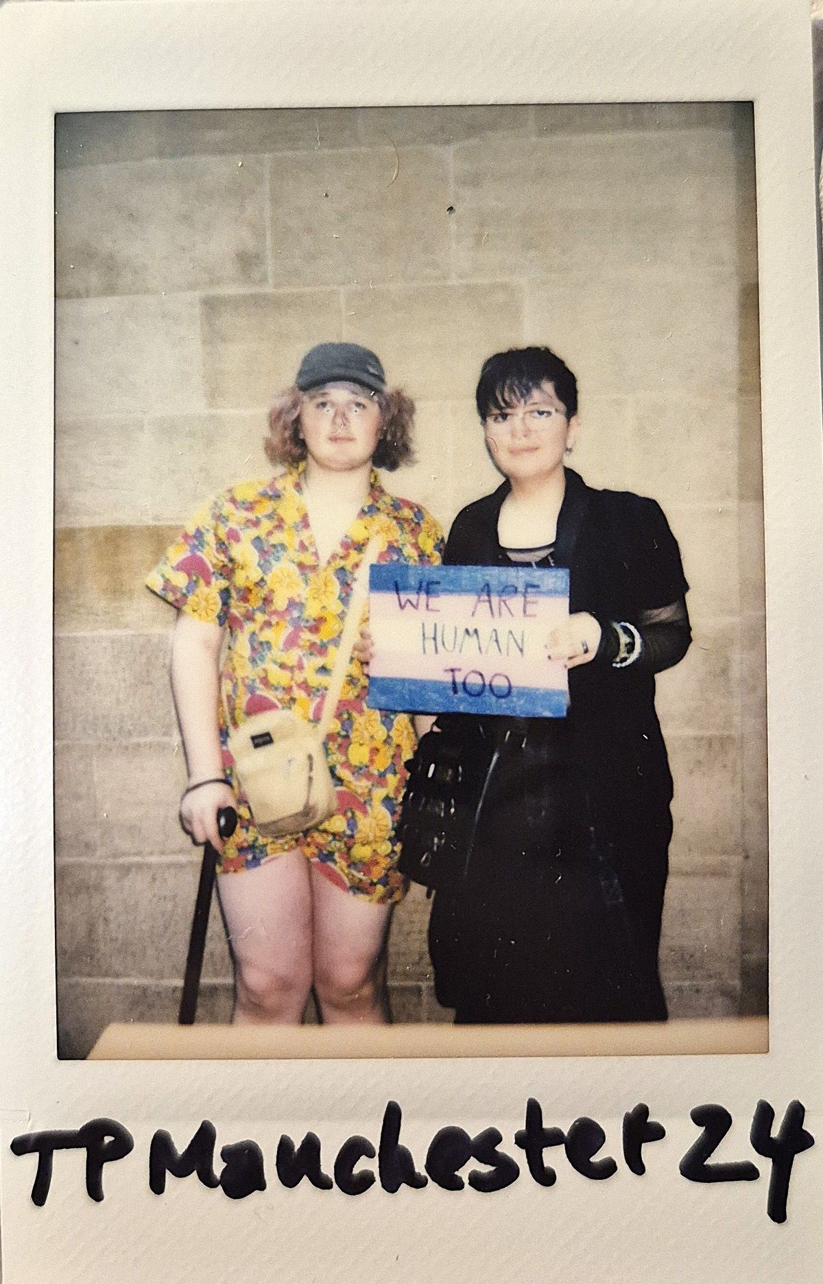 Two people stand together, one holding a sign saying “We Are Human Too.”