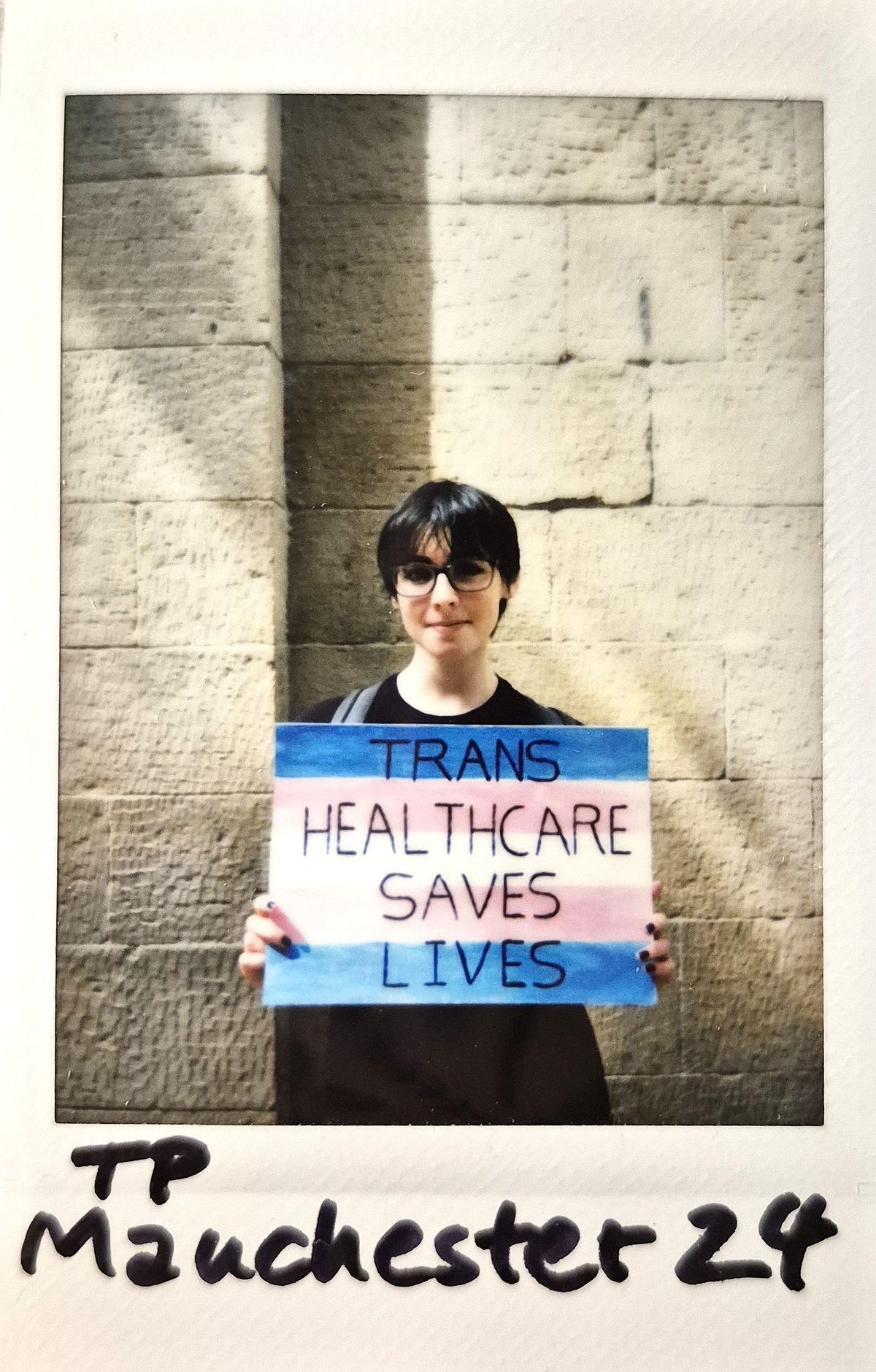 A person holding a colorful sign reads, "Trans healthcare saves lives," against a brick wall background.