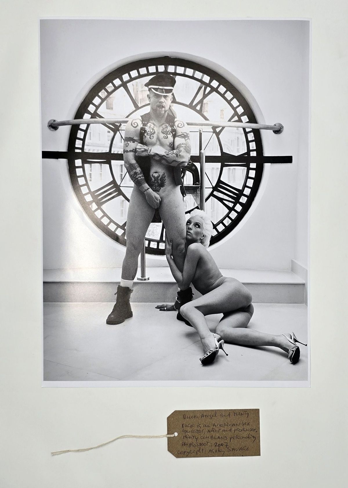 Black and white photograph of two people. One is stood up and only wearing a waistcoat and a hat, the other is on the floor leaning against the other and is naked.