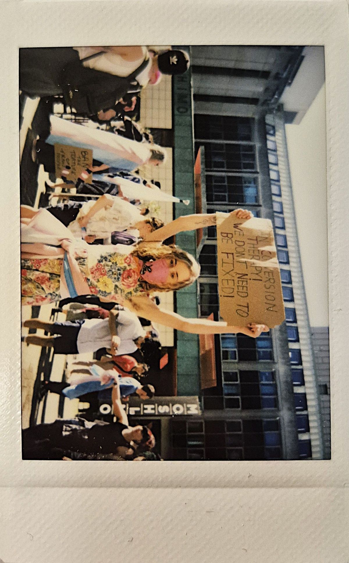 A person in a floral dress and mask holds a protest sign which says "BAN CONVERSION THERAPY WE DON'T NEED TO BE FIXED".