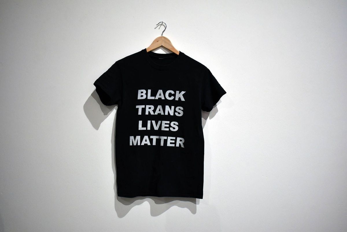 A black T-shirt hangs on a wooden hanger, with the text “Black Trans Lives Matter” in bold white text.