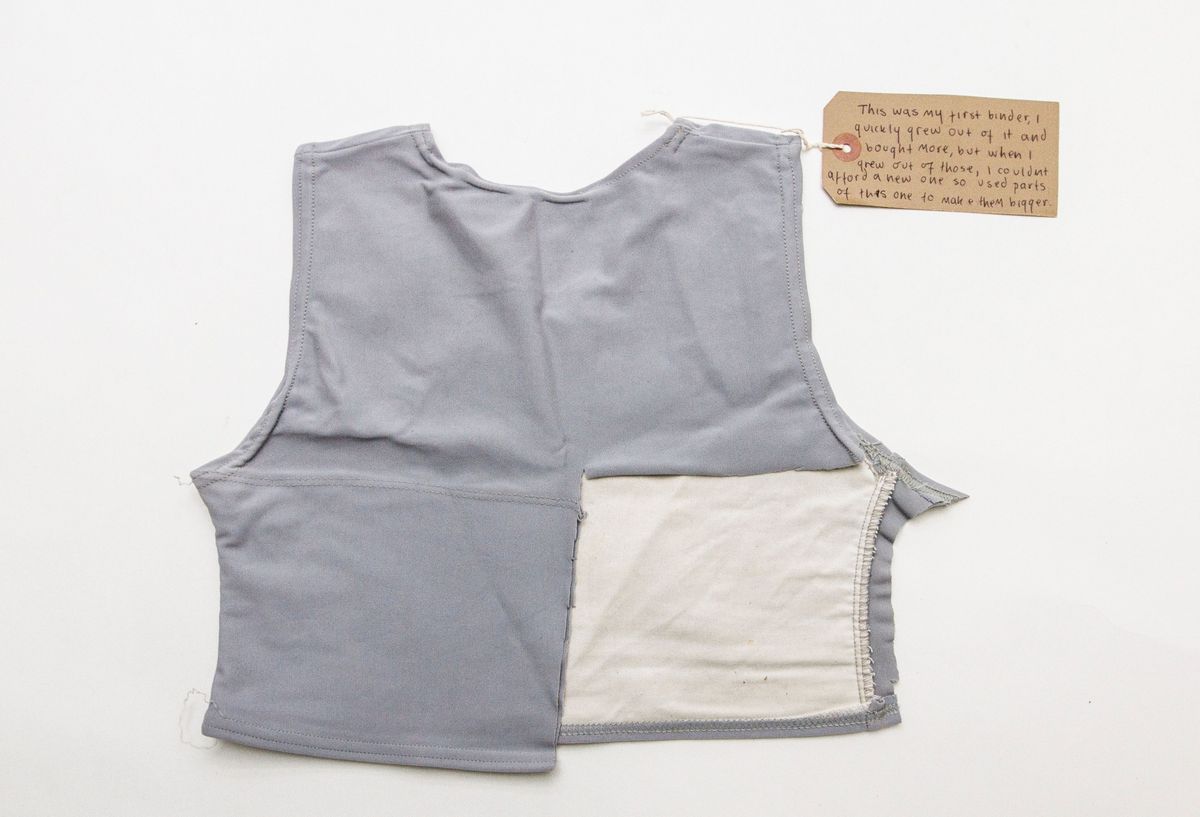 Grey chest binder displayed back-side-up on a white background. A square of fabric has been removed from the lower right side of the garment.