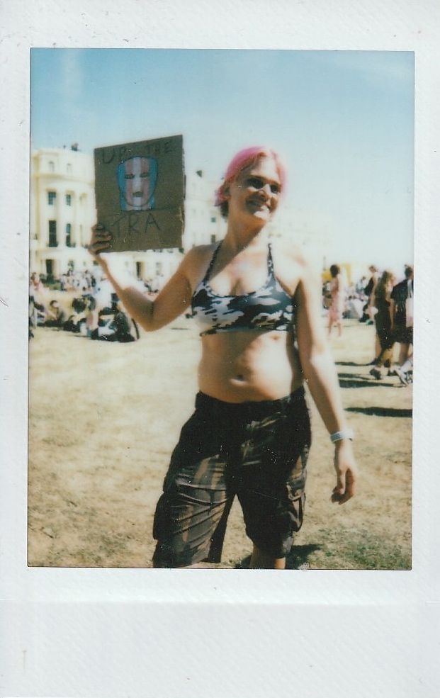 A person with pink hair holds a hand-drawn sign which says "UP THE TRA".