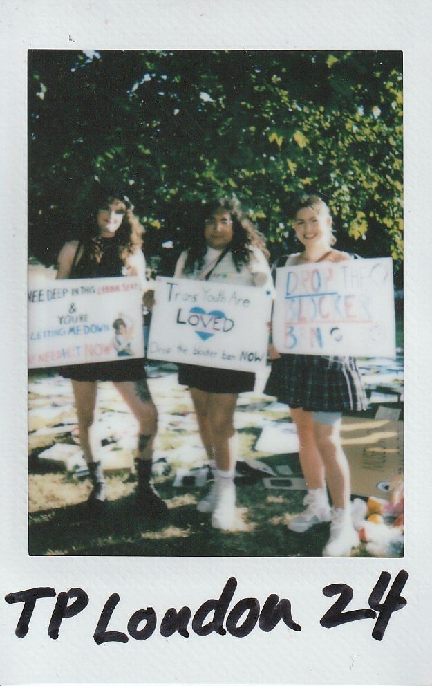 Three people stand together outdoors, holding signs supporting trans youth, under a tree with sunlight filtering.