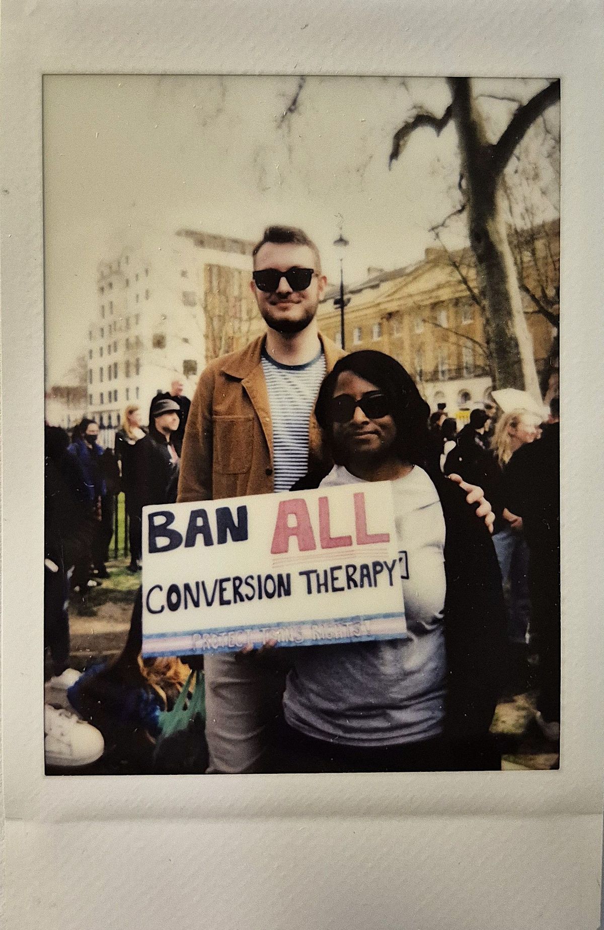 Two individuals at a public event hold a "Ban All Conversion Therapy" sign while standing closely together.