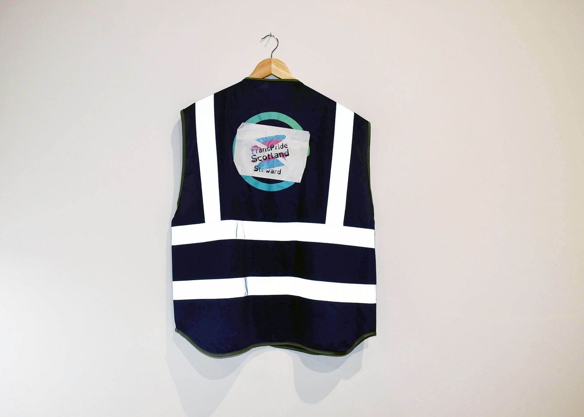 A dark vest with white reflective stripes, which says 'Trans Pride Scotland Steward' hangs from a wooden hanger on a plain wall.