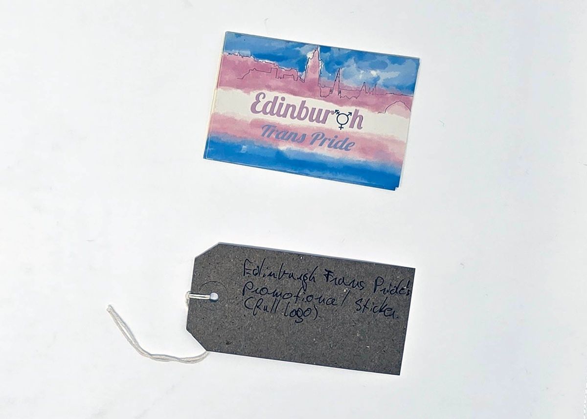 Rectangular sticker with the text "Edinburgh Trans Pride" (the 'g' in Edinburgh has been replaced with the trans symbol). The background is in the trans colours with a line drawing of a city skyline. With a brown, handwritten luggage tag.