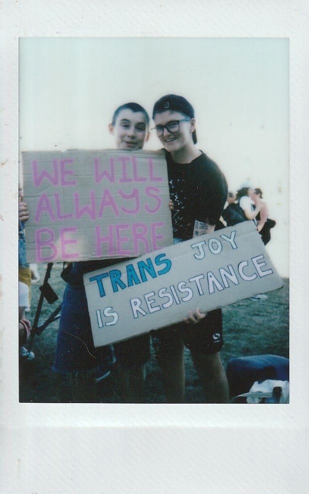 Two individuals holding signs read, "We will always be here" and "Trans joy is resistance."