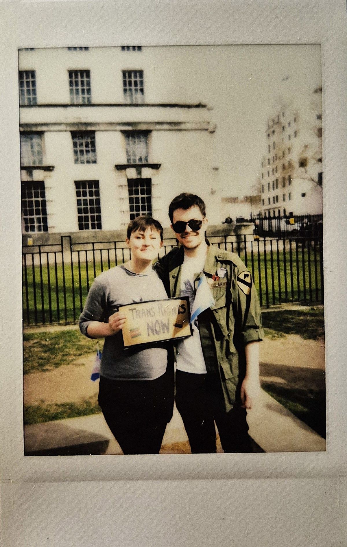 Two people stand together smiling, holding a sign which says "TRANS RIGHTS NOW".