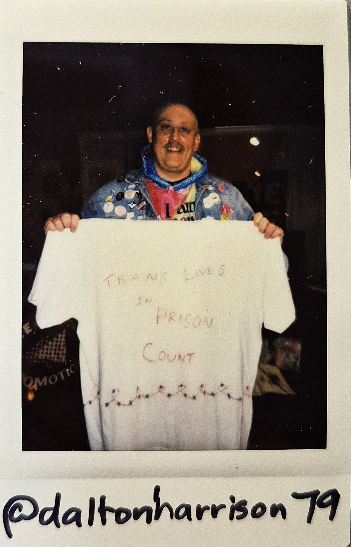 A person holding a shirt with the message "TRANS LIVES IN PRISON COUNT," smiling brightly.