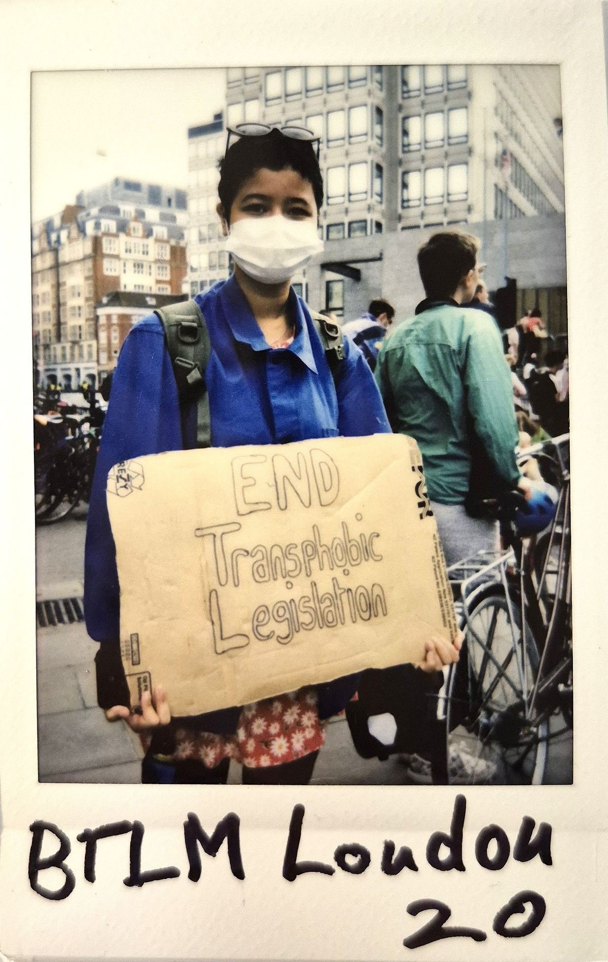 A person wearing a mask holds a sign that reads, "END Transphobic Legislation".