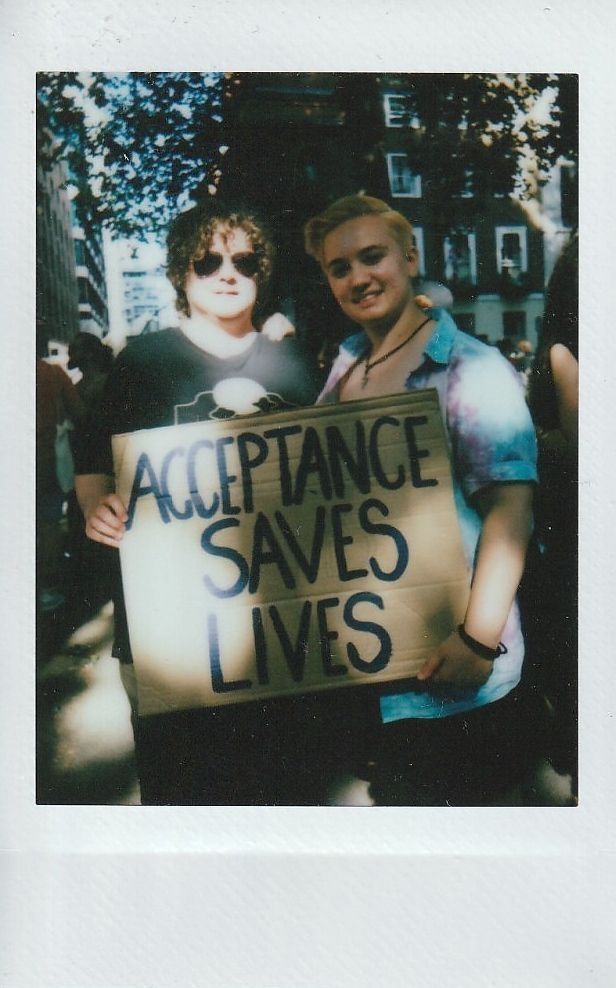 Two people are holding a cardboard sign that says "Acceptance Saves Lives" in a sunny outdoor setting.