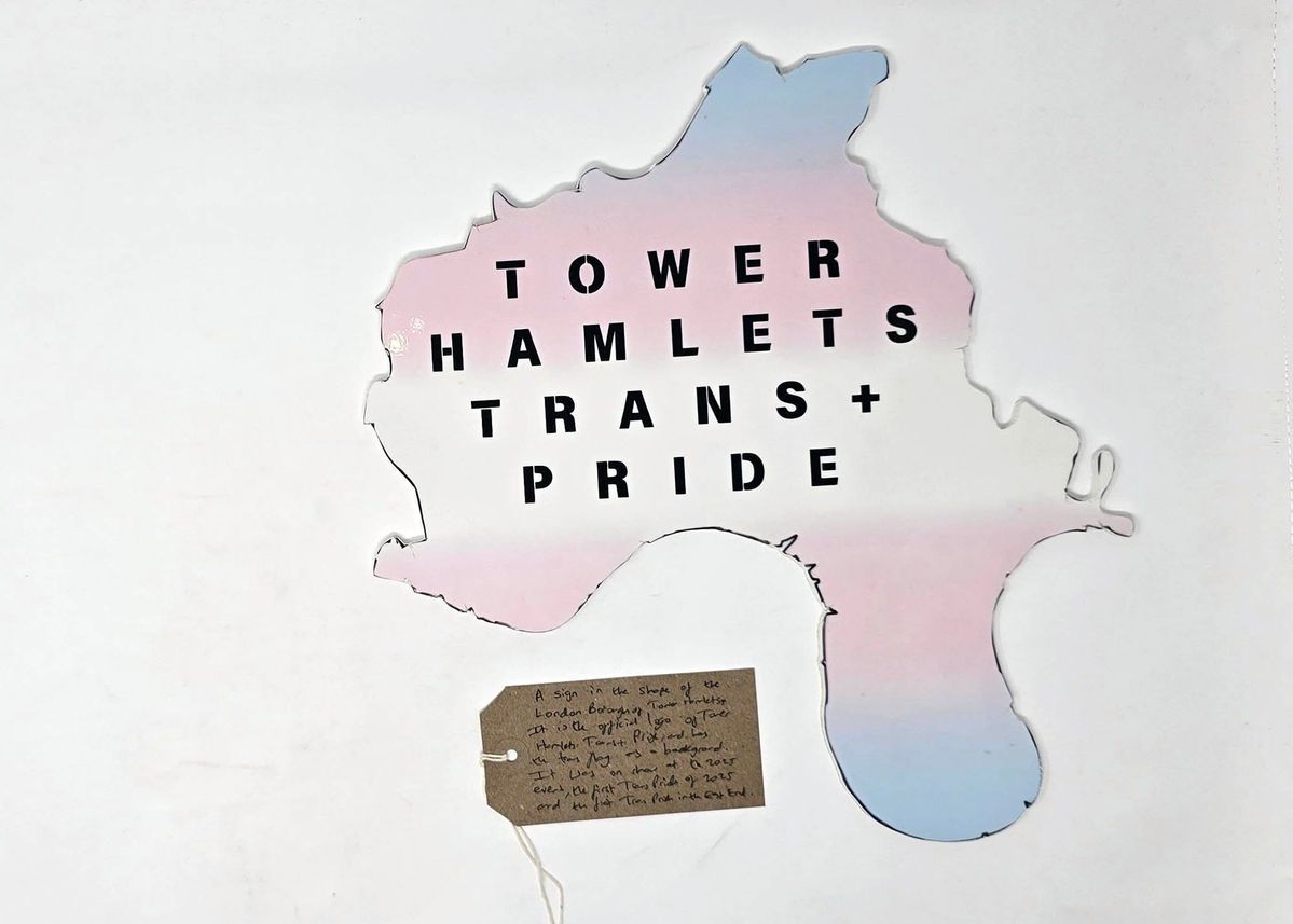 A foam board Tower Hamlets Trans+ Pride logo - which is the shape of the boundary of the London Borough of Tower Hamlets. It is colored in the blue, white and pink trans pride colours. With a brown, handwritten luggage tag.