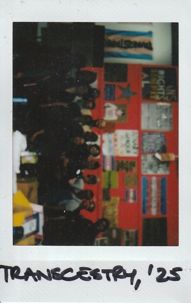 A group of people gathers in front of a colorful wall filled with various posters and signs.