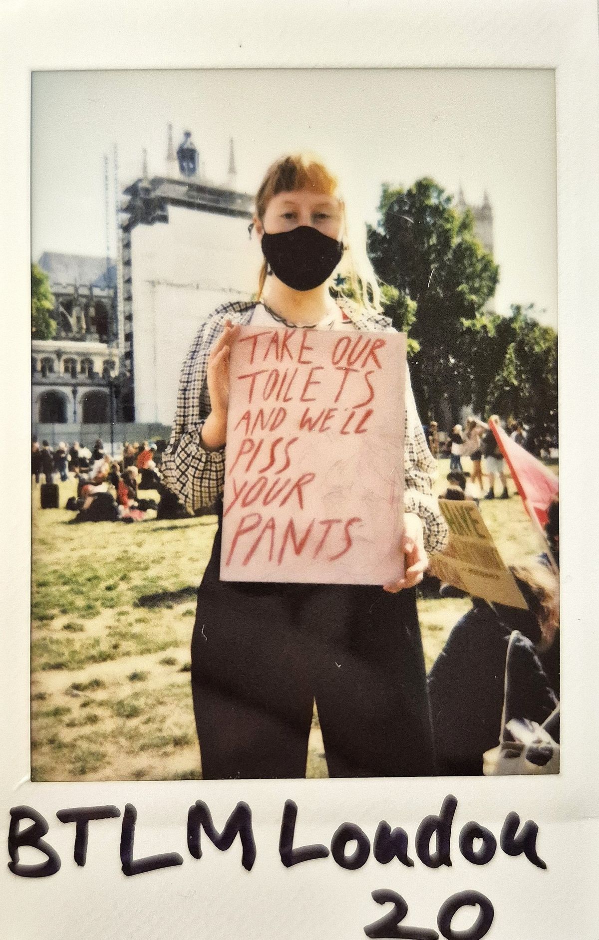 A person wearing a mask holds a sign in a park, protesting about toilet access rights for trans people.