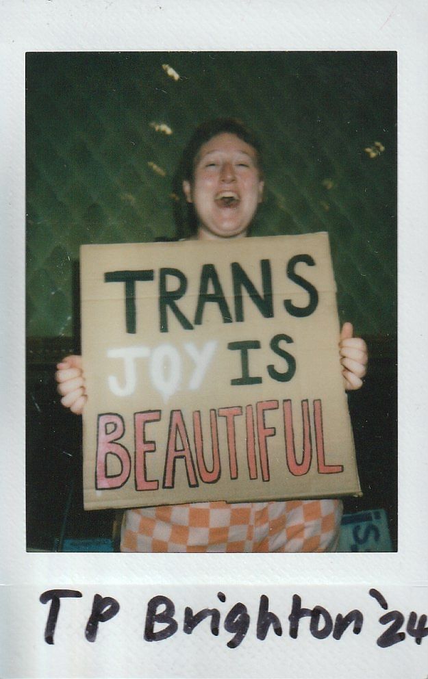 A person holds a sign reading "TRANS JOY IS BEAUTIFUL" while smiling joyfully.