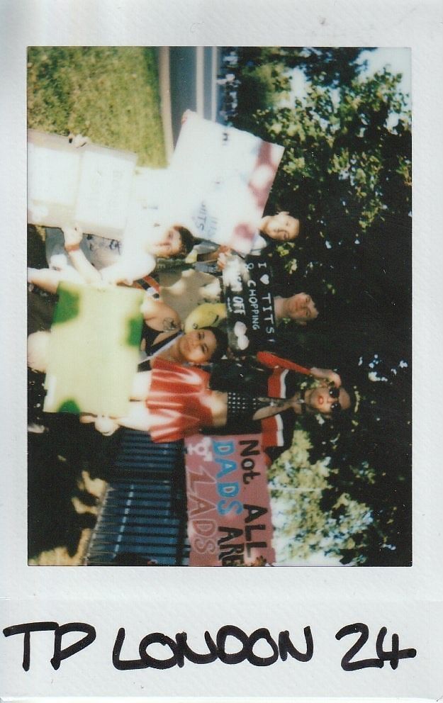 A group of people hold colorful signs outdoors, with trees visible in the background.