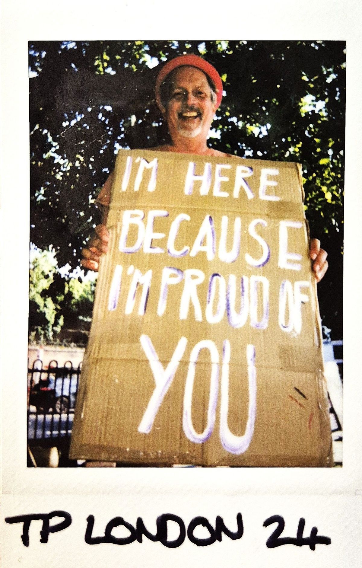 A smiling person holds a cardboard sign that says, "I'm here because I'm proud of you."