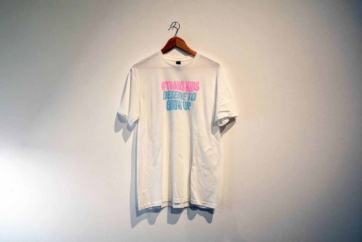 A white T-shirt with colorful text hangs on a wall emphasizing a supportive message.