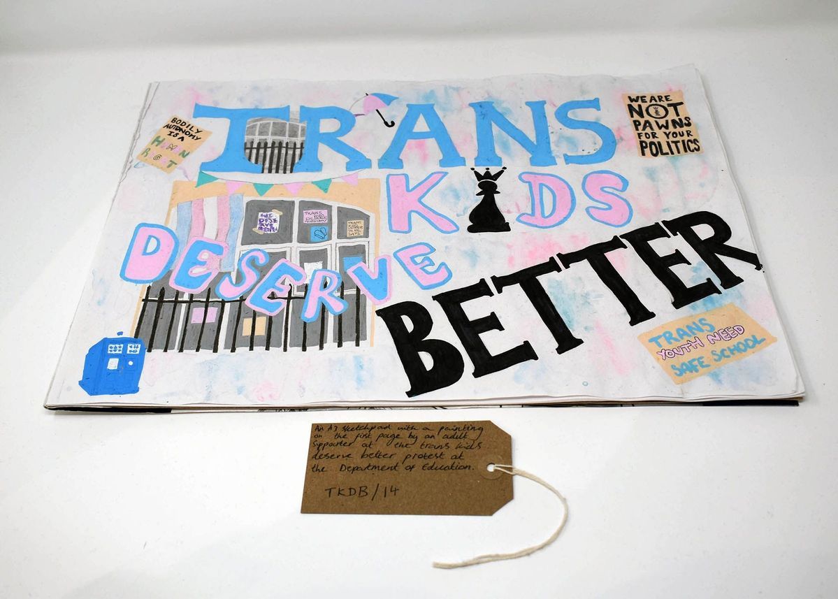 An A3 sketch pad with a hand painted Trans Kids Deserve Better poster. It features smaller paintings of other placards and scenes from the Department for Education Occupation/Encampment and a blue tardis. With a brown, handwritten luggage tag.