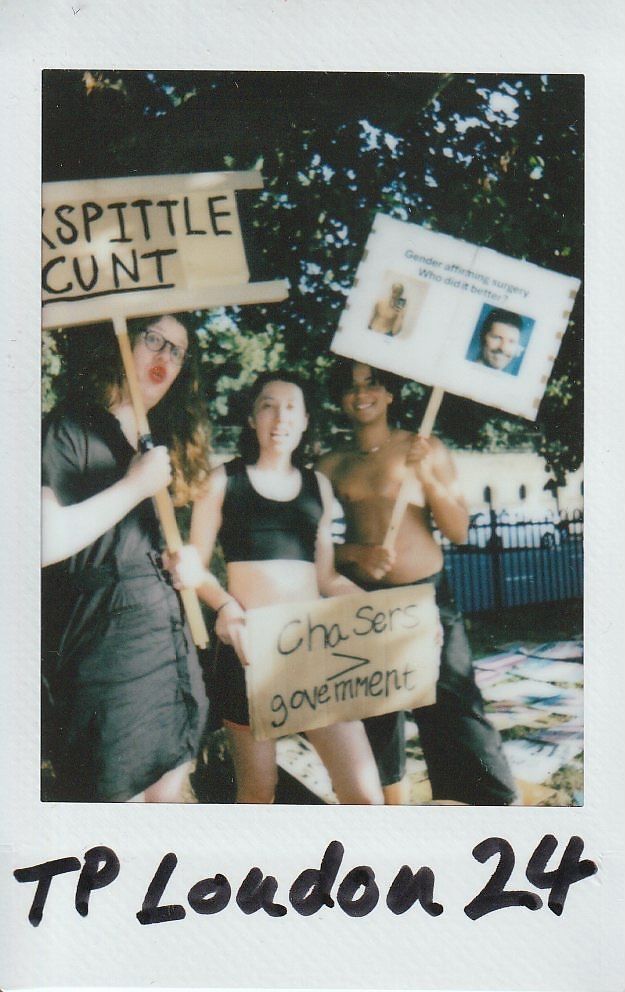 Three people hold protest signs, expressing opinions on gender and government, outdoors in a sunlit setting.